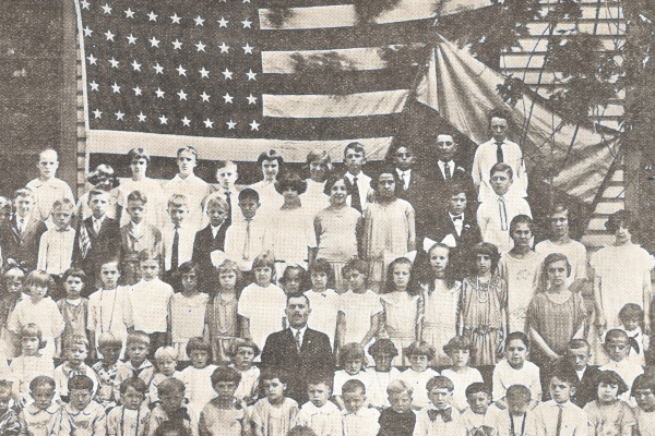 Group photo of early 20th century children and adults in front of a large US flag and a smaller Ukrainian flag